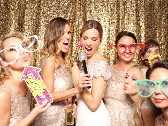 Bridal party laughing and posing with photo booth props in front of a gold sequin backdrop.
